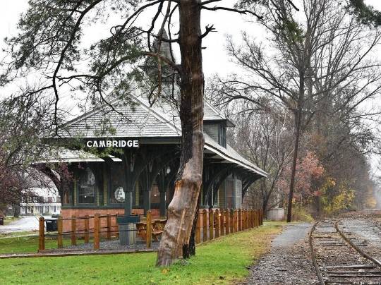 Cambridge New York Train Station. Photograph of an old wooden and brick train station labeled ‘CAMBRIDGE’, with a wooden platform and a large bare tree in the foreground. Train tracks run alongside the platform, extending into the distance. The background is filled with trees and shrubs displaying autumn colors.