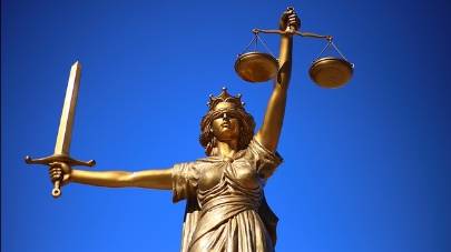 Photograph of a bronze statue of Lady Justice, depicted in classical style with a flowing robe and a crown. She is holding a sword in her right hand and a set of scales in her left hand, symbolizing the power and balance of justice. The statue is set against a clear blue sky.