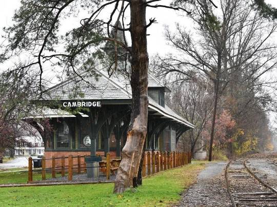 Cambridge New York Train Station. Photograph of an old wooden and brick train station labeled ‘CAMBRIDGE’, with a wooden platform and a large bare tree in the foreground. Train tracks run alongside the platform, extending into the distance. The background is filled with trees and shrubs displaying autumn colors.