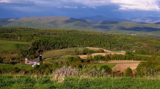 Photograph of a scenic view of a lush green valley with a few buildings and a silo, set against a backdrop of mountains in various shades of green and brown. The sky above is clear and blue with a few scattered clouds.