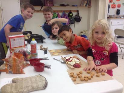 Photograph of three children engaged in a craft project at a table covered with a white tablecloth. The table is strewn with construction paper, glue, and other craft supplies. A red bowl and a bag of chips are also visible on the table. The background appears to be a kitchen or classroom, with cabinets and a sink.