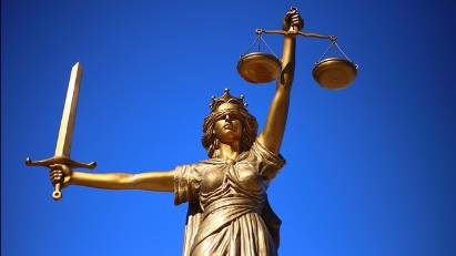 Photograph of a bronze statue of Lady Justice, depicted in classical style with a flowing robe and a crown. She is holding a sword in her right hand and a set of scales in her left hand, symbolizing the power and balance of justice. The statue is set against a clear blue sky.