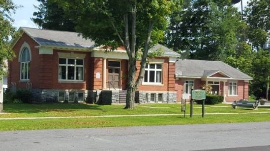 Photograph of a red brick building labeled ‘The Village of Cambridge Library’, with a green lawn in the foreground and trees in the background. The building features a gray slate roof and white trim around the windows, and is located on a street with a sidewalk and a streetlamp.