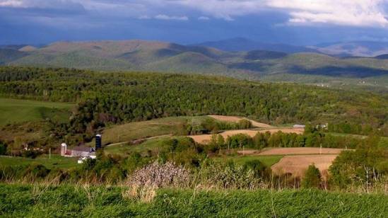 Photograph of a scenic view of a lush green valley with a few buildings and a silo, set against a backdrop of mountains in various shades of green and brown. The sky above is clear and blue with a few scattered clouds.