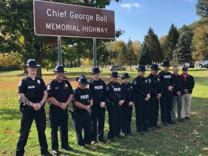 Photograph of a group of police officers in dark uniforms and hats standing in front of a brown sign that reads ‘Chief George Bell Memorial Highway’, located in a grassy area with trees in the background.