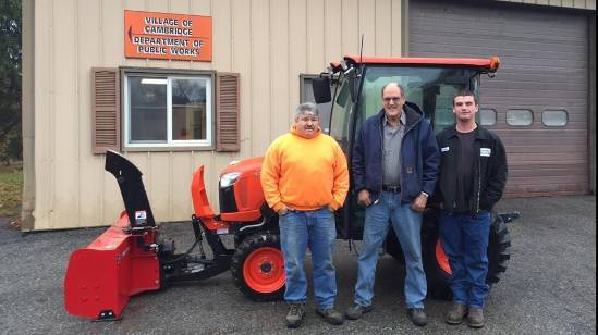 Photograph of three individuals in work attire standing in front of a red Kubota tractor with a snow blower attachment, located at the ‘Village of Cambridge Department of Public Works’. The building sign is visible in the background.