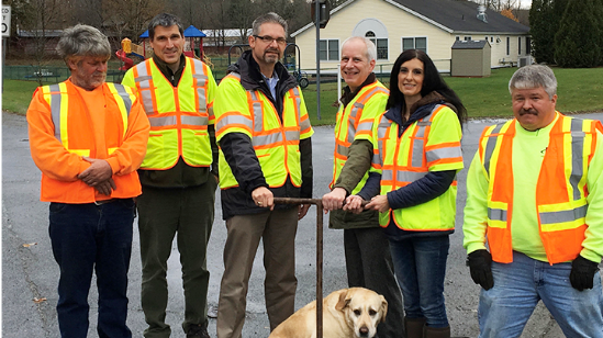 Photograph of a group of people wearing orange safety vests, standing in front of a white building with a gray roof. A dog is sitting on the ground in front of the group. The sky is gray and overcast.