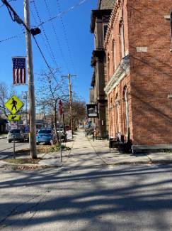 Village of Cambridge, NY corner of Main Street and Washington Street with a view of a red brick building with a sidewalk in front of it. There are a few cars parked along the street and an American flag hanging from a pole.